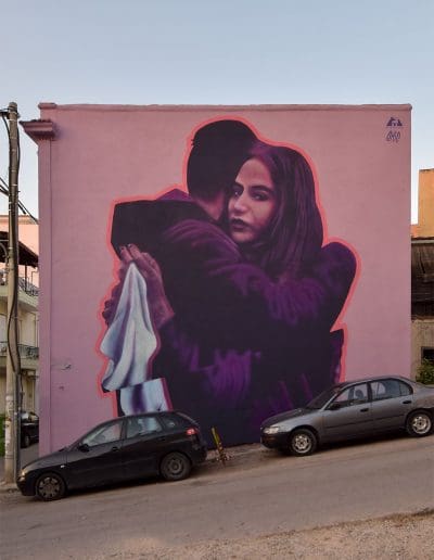 Mural by emic in Patras, Greece featuring two figures hugging with the female gazing out to the viewer as she holds a white cloth to represent peace
